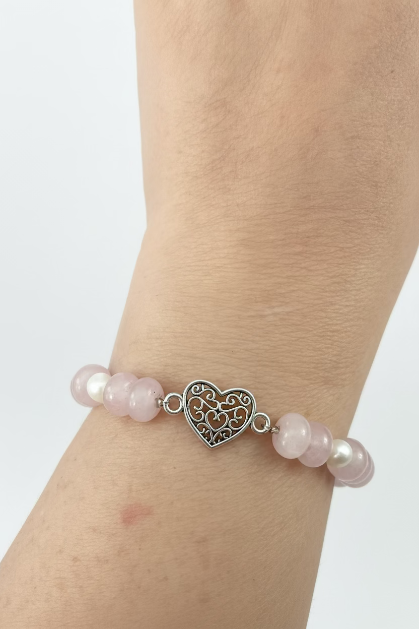 A close-up of a person's wrist wearing a beaded bracelet with light pink and white pearls, a silver filigree heart charm, and a pink stone bead.