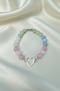 A beaded bracelet with alternating rows of pale green, white, light blue, and pink round beads, featuring a central silver-tone open heart charm, displayed on a light wood background.