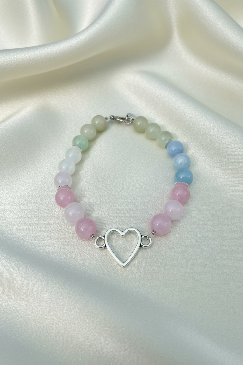 A beaded bracelet with alternating rows of pale green, white, light blue, and pink round beads, featuring a central silver-tone open heart charm, displayed on a light wood background.