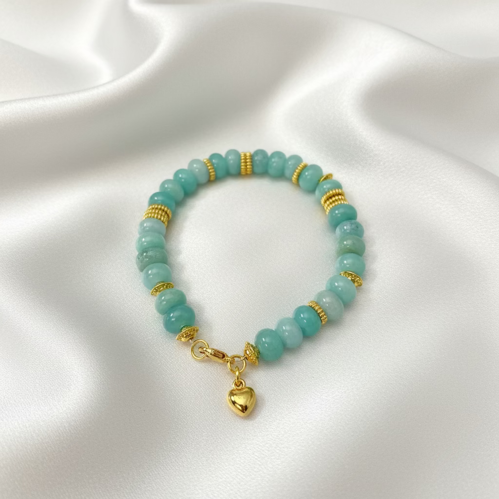 A beaded bracelet made of light aqua blue-green round beads separated by small gold spacer rings, with a gold clasp and dangling heart charm, displayed on a dark textured surface.