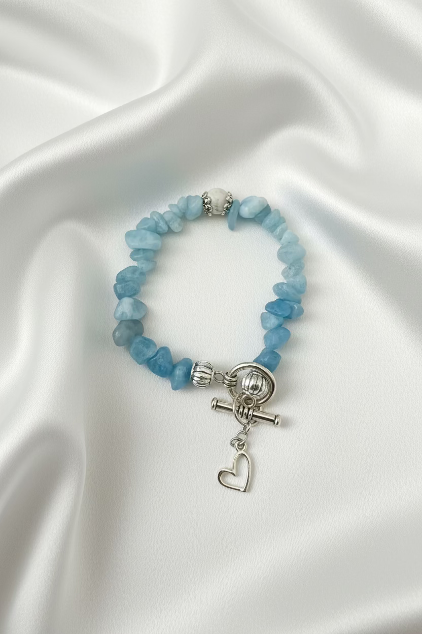 A beaded stretch bracelet made of light blue stone chips with a silver toggle clasp and dangling heart charm, displayed on a dark textured surface.