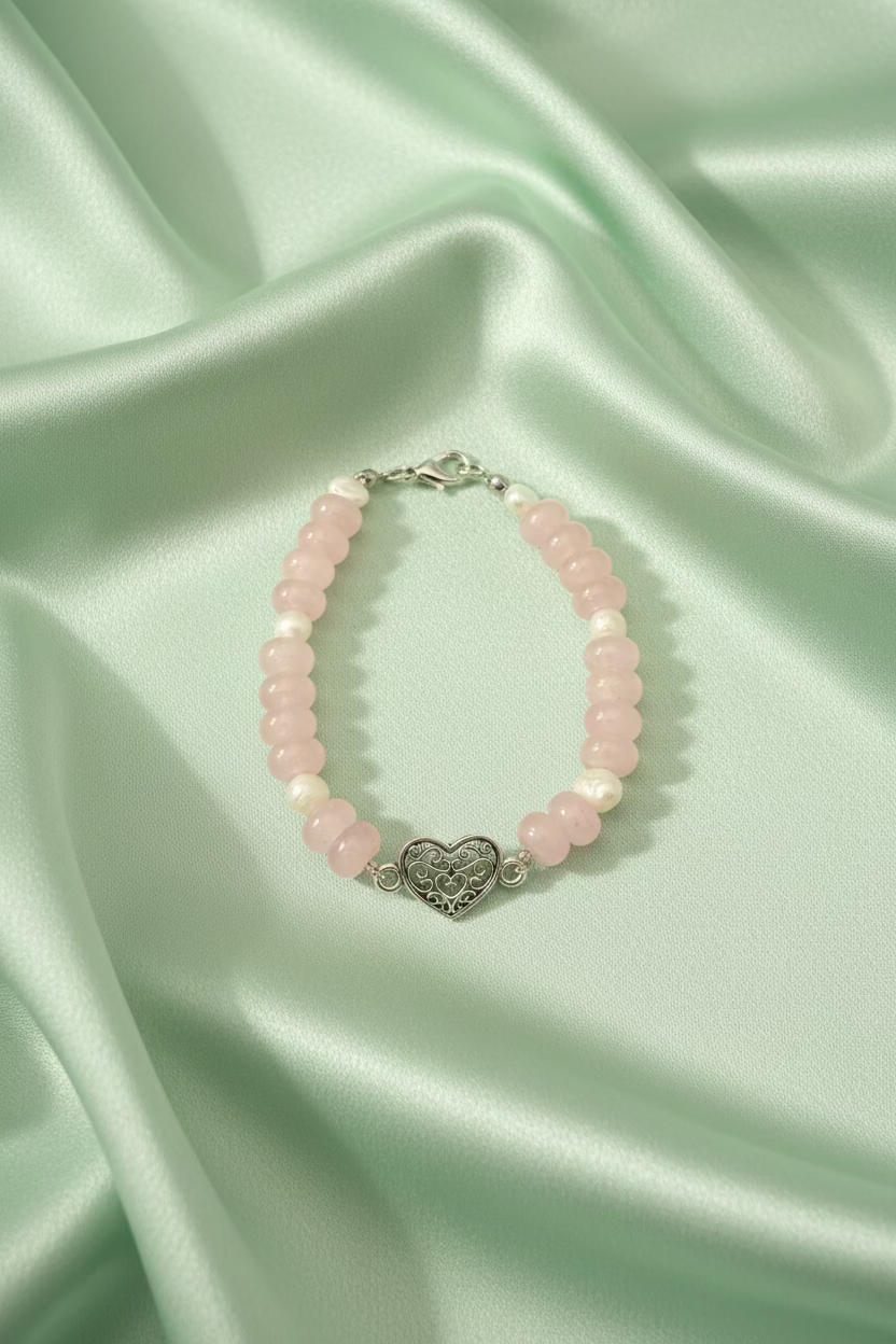 A beaded bracelet featuring light pink rose quartz beads, small white pearl accents, and a central silver filigree heart charm, displayed on a dark textured surface.