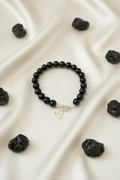 A black beaded bracelet with a silver lobster clasp and a dangling open heart charm, displayed on a dark textured surface.