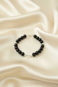 A beaded bracelet featuring glossy black round beads, a few silver-toned metallic beads, and a small silver heart-shaped charm in the center, displayed on a dark textured surface.