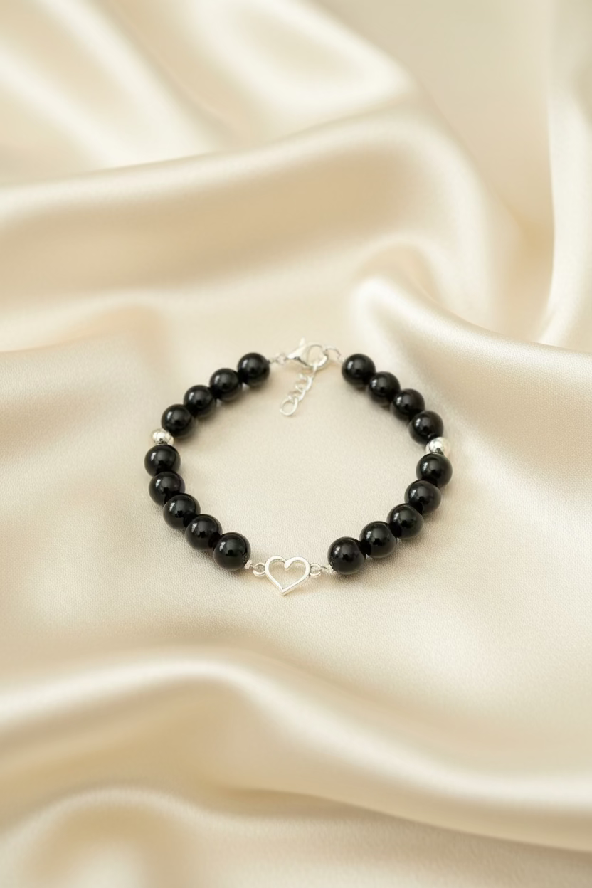 A beaded bracelet featuring glossy black round beads, a few silver-toned metallic beads, and a small silver heart-shaped charm in the center, displayed on a dark textured surface.