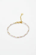 A delicate beaded bracelet with small faceted beads in pale pink, yellow, and white, accented by a gold-tone clasp and extender chain, displayed on a dark textured background.