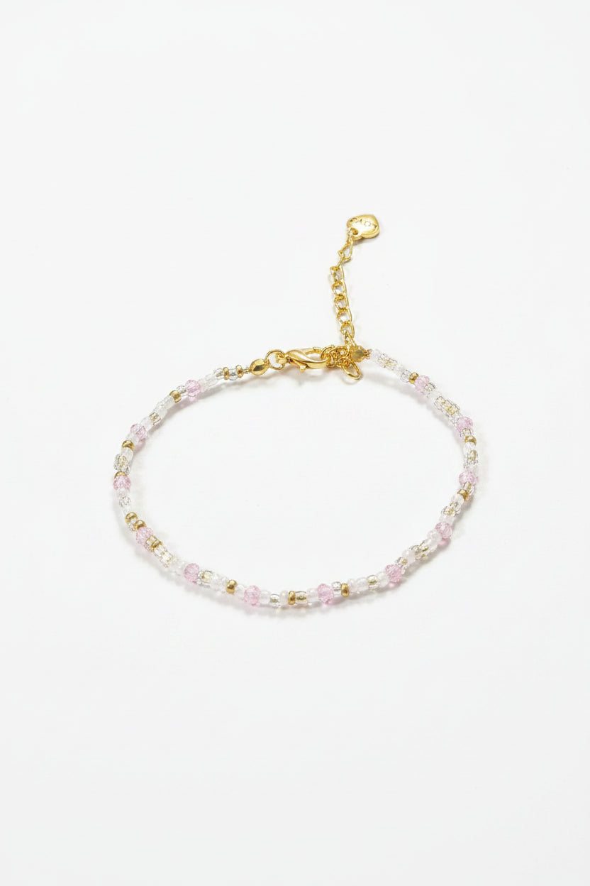 A delicate beaded bracelet with small faceted beads in pale pink, yellow, and white, accented by a gold-tone clasp and extender chain, displayed on a dark textured background.