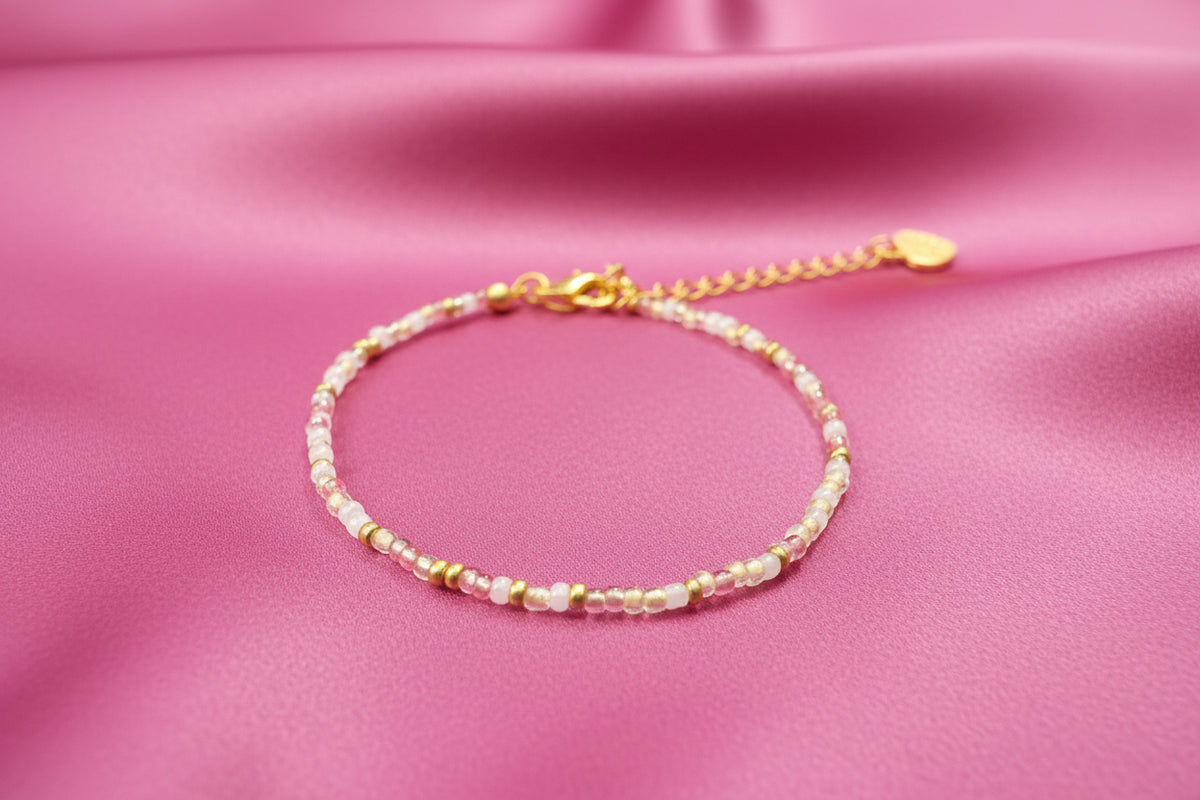 A beaded bracelet with small round beads in pink, gold, and white, featuring a gold chain extender and clasp, displayed on a textured black background.