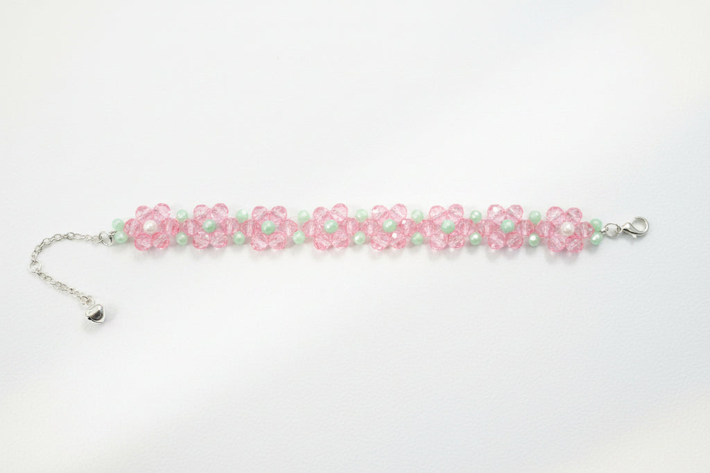 A handmade beaded bracelet featuring pink flower motifs with white centers, accented by light green beads and a silver-tone chain extender with a small heart charm, displayed on a dark textured background.