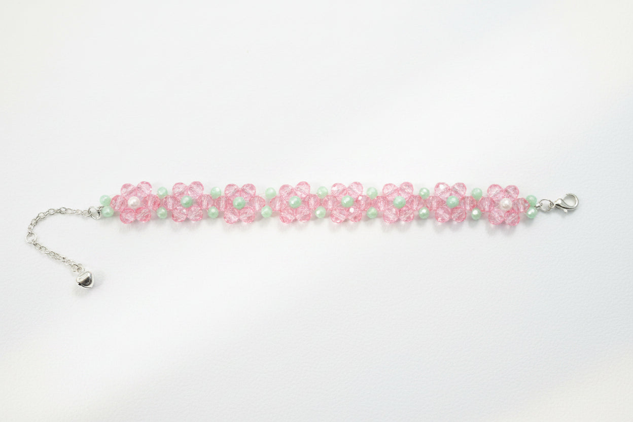 A handmade beaded bracelet featuring pink flower motifs with white centers, accented by light green beads and a silver-tone chain extender with a small heart charm, displayed on a dark textured background.