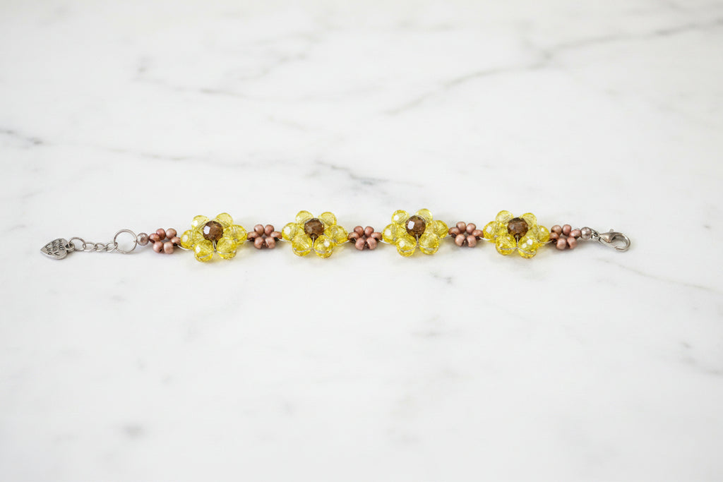 A handmade beaded bracelet featuring three yellow flower motifs with brown centers, accented by small pinkish-brown beads, displayed on a dark textured background.