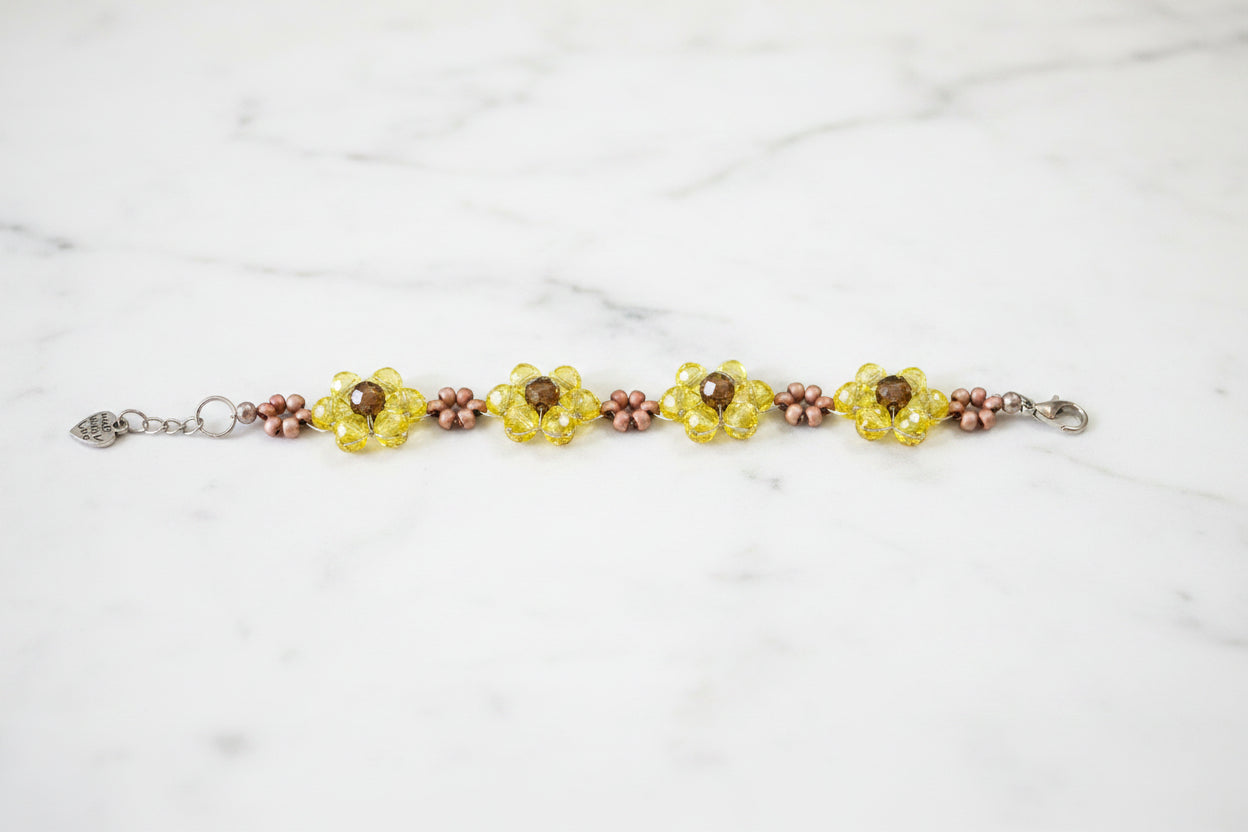 A handmade beaded bracelet featuring three yellow flower motifs with brown centers, accented by small pinkish-brown beads, displayed on a dark textured background.