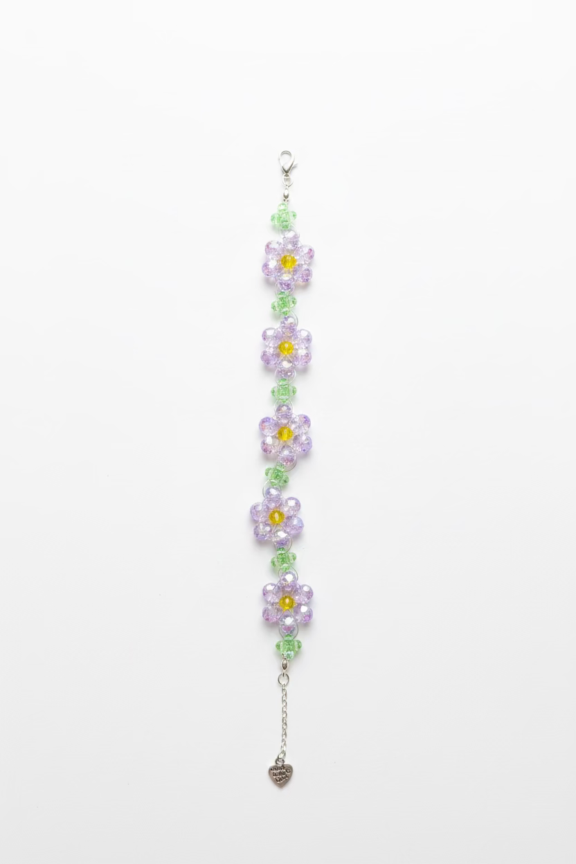 A handmade beaded bracelet featuring purple and green flower charms with yellow centers, attached to a silver lobster clasp and extension chain, displayed on a black fabric surface next to a laptop.
